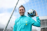 A woman smiles while posing for a photo and holding a soccer ball on her shoulder.