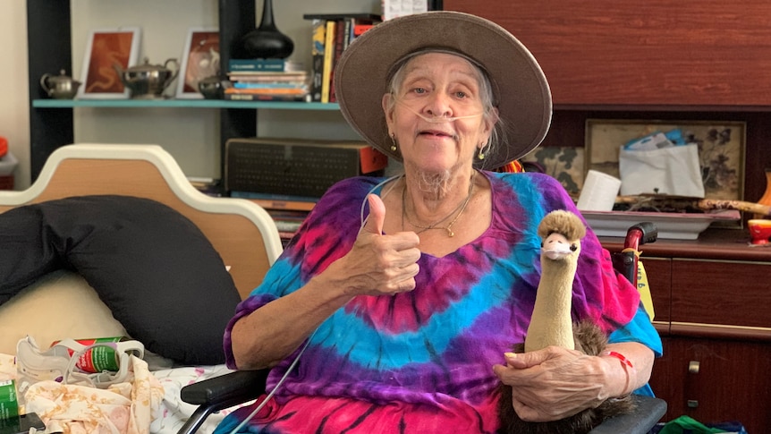 older woman of indigenous background wearing akubra hat and tie dye pink and blue shirt giving thumbs up while holding plush emu