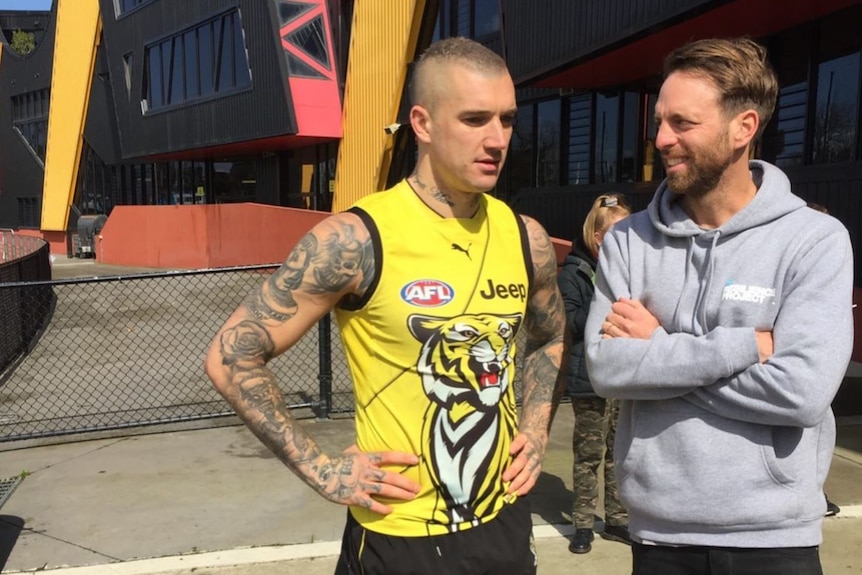 A richmond football player stands next to a man with a grey hoodie and his arms crossed 