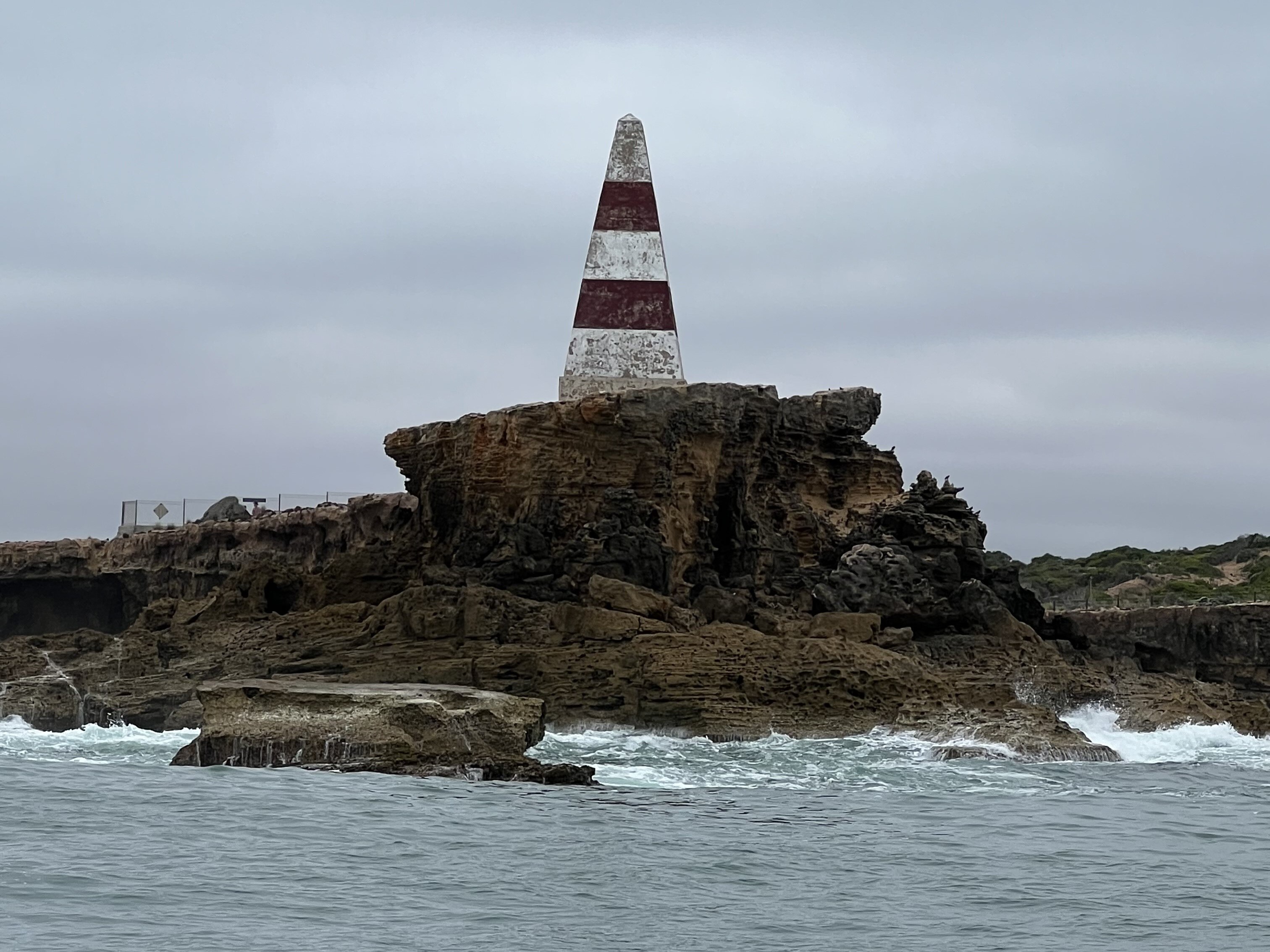 A red and white pyramid shaped obelisk atop a rocky cliff with water below.