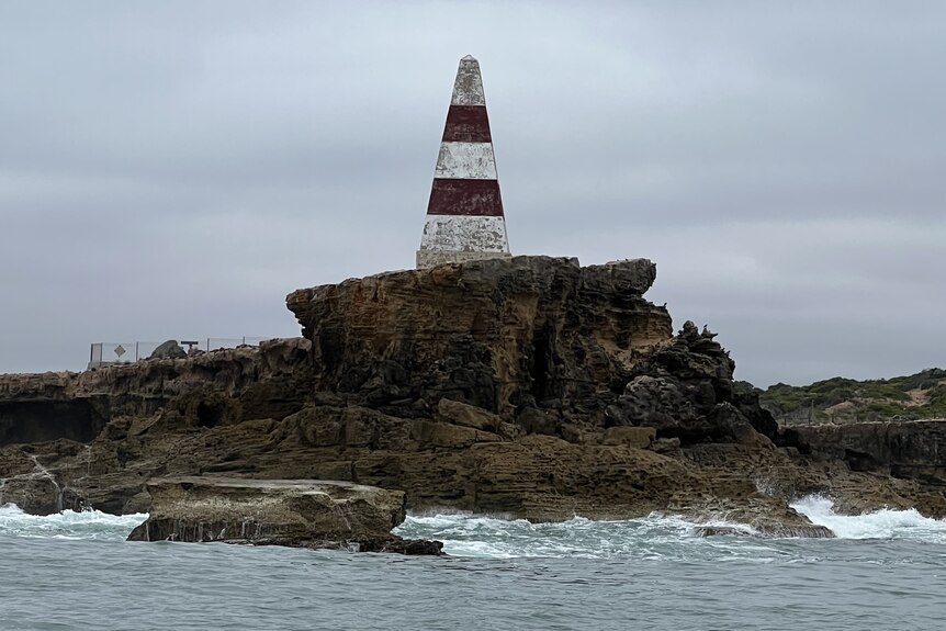 A red and white pyramid shaped obelisk atop a rocky cliff with water below.