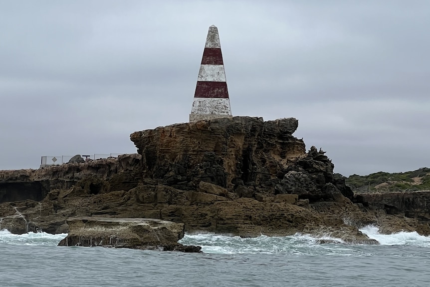 A red and white pyramid shaped obelisk atop a rocky cliff with water below.