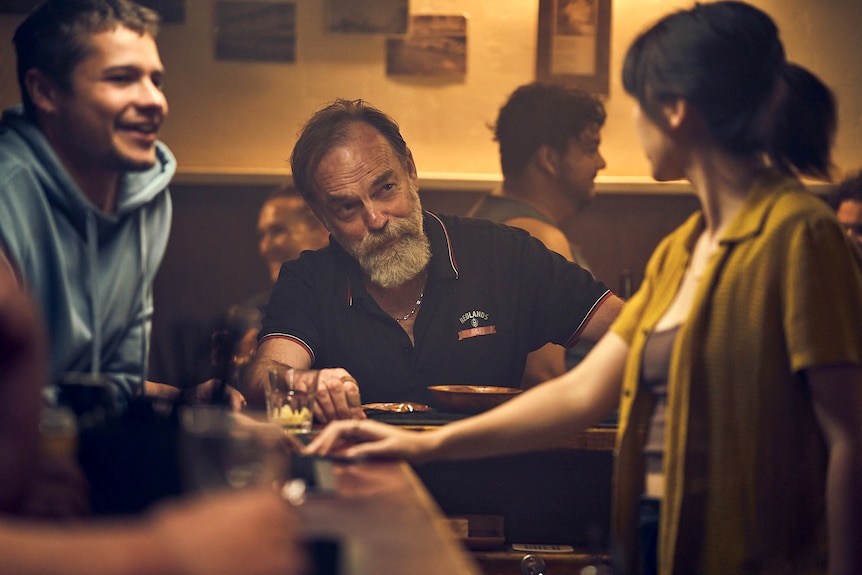 Hugo Weaving, in character, leans on the bar of a pub, chatting to a young man and a young woman behind the bar.