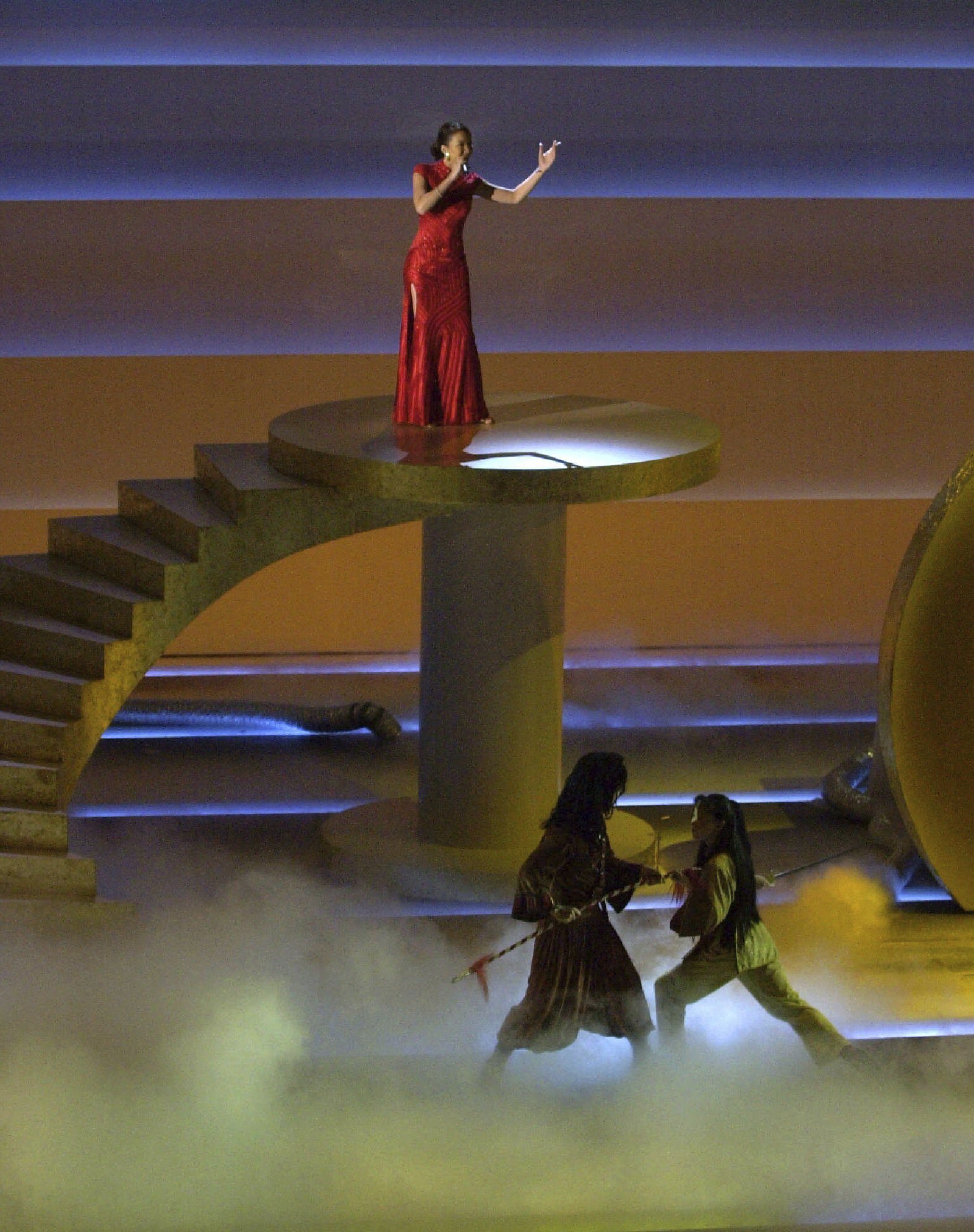 Woman in red gown gestures as she sings into microphone on raised platform.