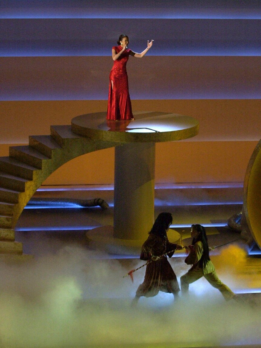 Woman in red gown gestures as she sings into microphone on raised platform.
