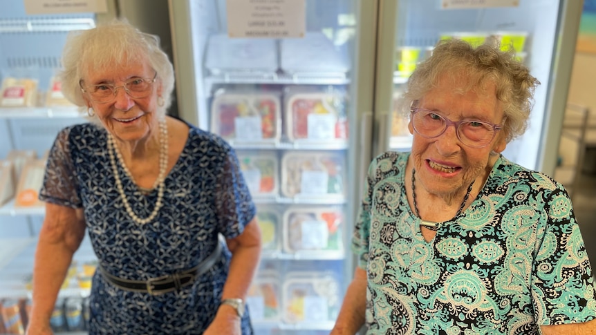 two elderly women with patterned dresses stand in front of a fridge stocked with meals