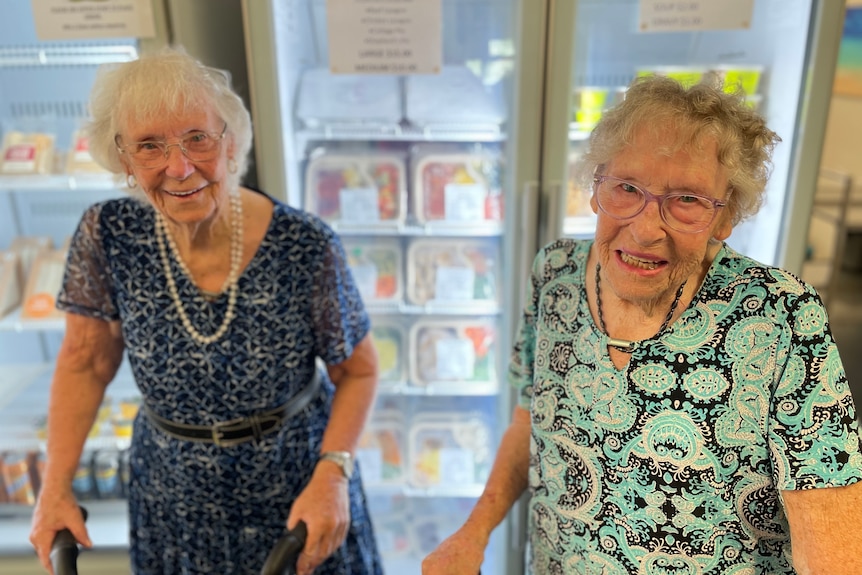 two elderly women with patterned dresses stand in front of a fridge stocked with meals