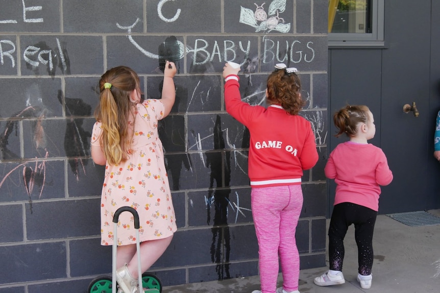 Three young children draw with chalk on a grey brick wall