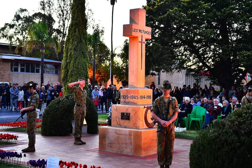 Two young army cadets stand next two a cross war memorial while another salutes while a crowd watches.