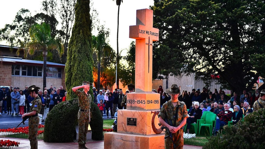 Two young army cadets stand next to a war memorial and another salutes while a crowd watches.