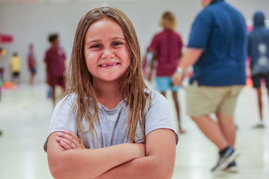 Young girl with long hair, smiling, wearing t-shirt and arms crossed, though looking happy.