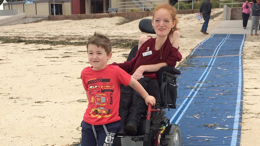 boy on front of lady's wheelchair on mat at beach