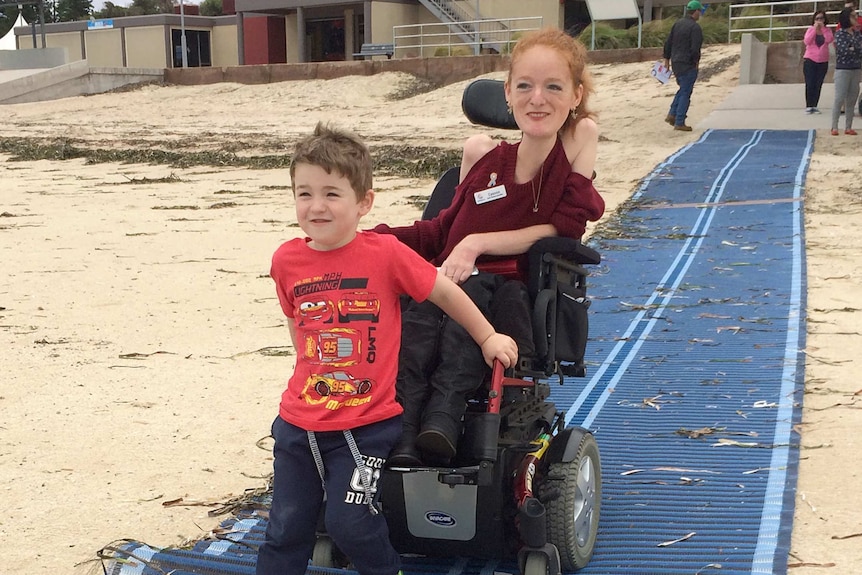 boy on front of lady's wheelchair on mat at beach