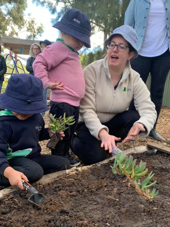 A woman kneeling next to a garden bed gestures and explains something to a toddler standing next to her.