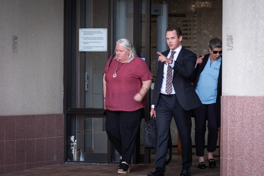 Two women and a man walk through the front automatic doors of a beige building