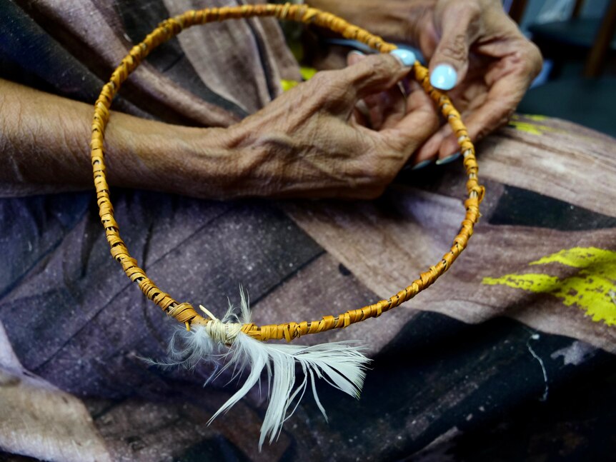 A woven halo shaped head dress with a cockatoo feather is held in a pair of hands