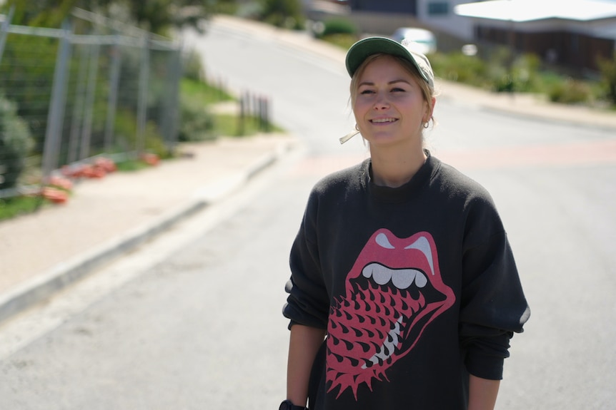 A blonde woman wearing a cap and black sweatshirt smiles standing on a suburban road