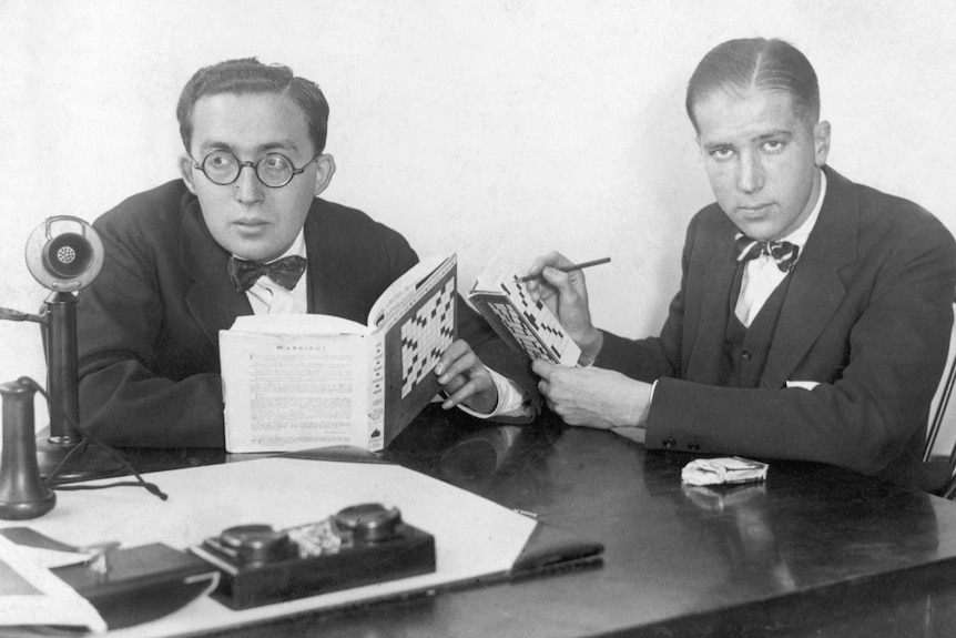 Black and white photo of two serious-looking men at a table, both wearing suits and holding crossword books.