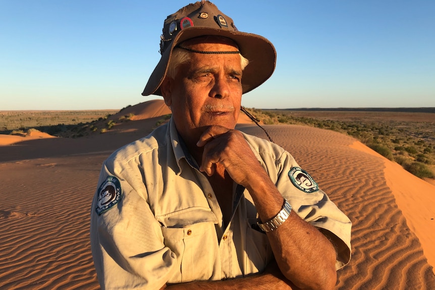 A ranger stands on a sand dune in the desert, looking into the distance with his hand on his chin.