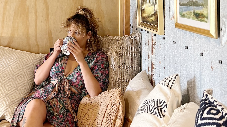 A woman sips from a mug while sitting on a couch with legs crossed, surrounded by cushions and Australian landscape art.