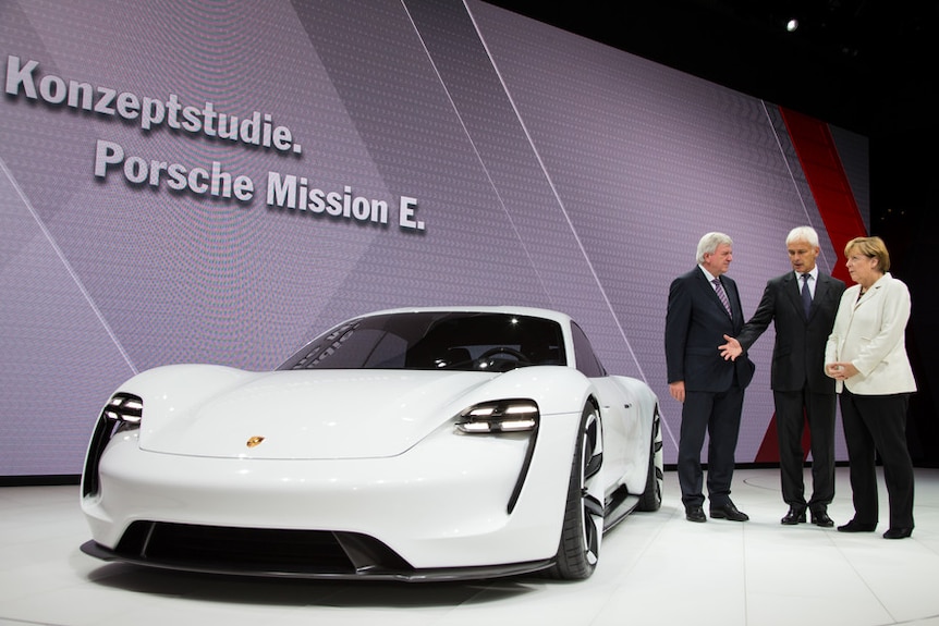 A white Porsche sports car in a display room, with three people standing nearby.