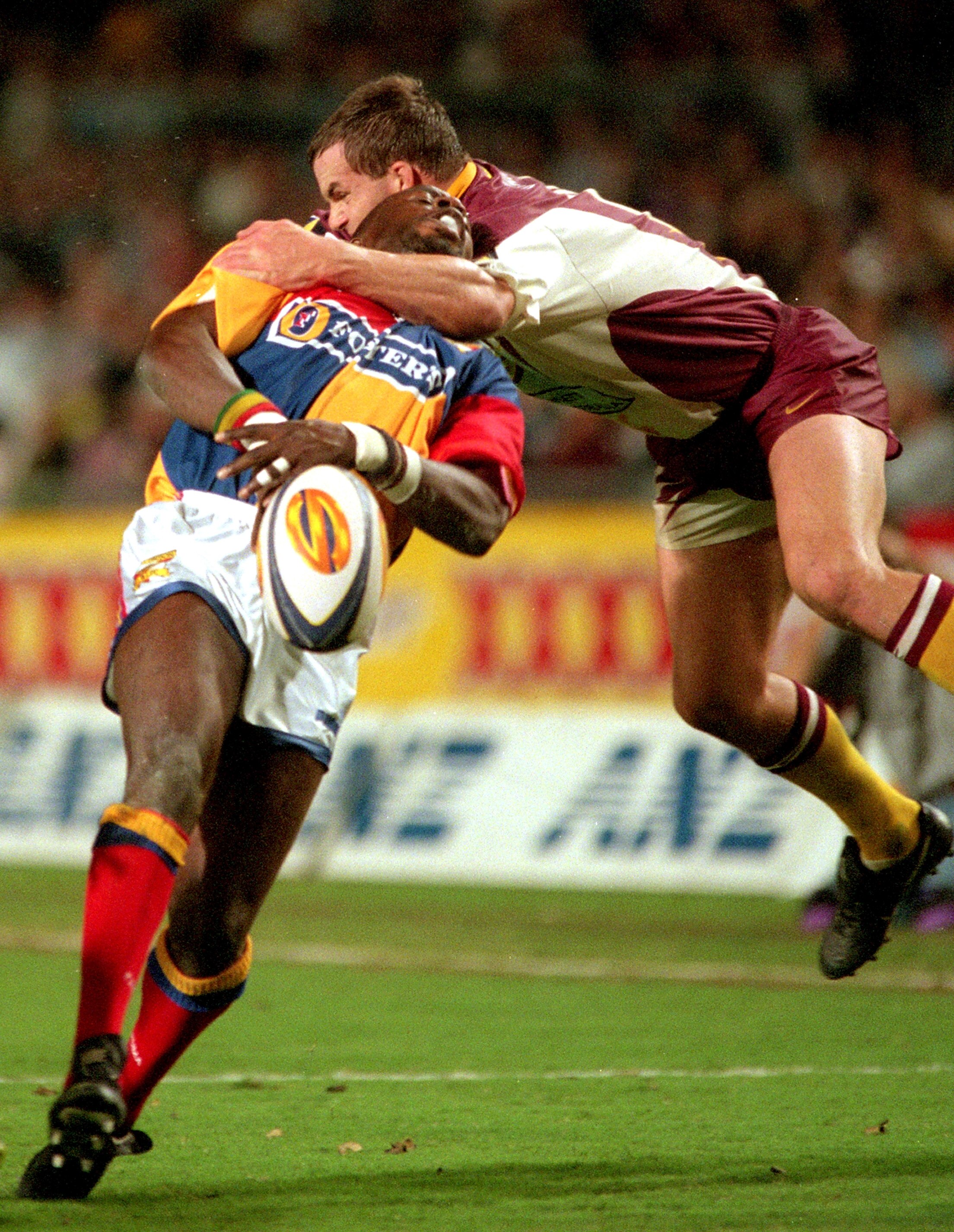Martin Offiah drops the ball while getting hit in the throat by a jumping Brisbane Broncos player during a rugby league game.