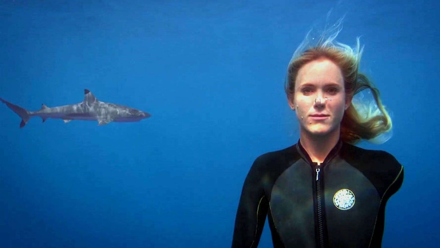 A woman with one arm wears a wet suit underwater as a shark swims in the background.