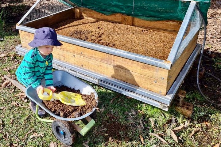 A child in a stripy shirt and hat uses a spade and wheelbarrow to move compost