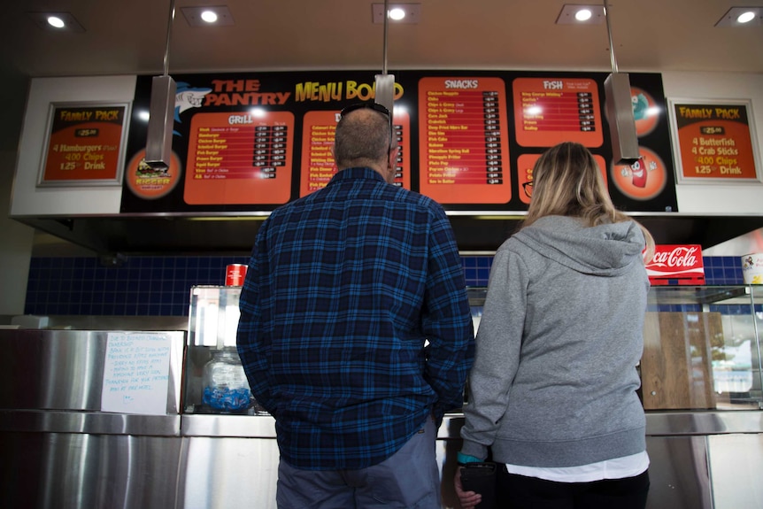 a low angle shot. A man and woman stand in front of a takeaway counter looking at the menu.