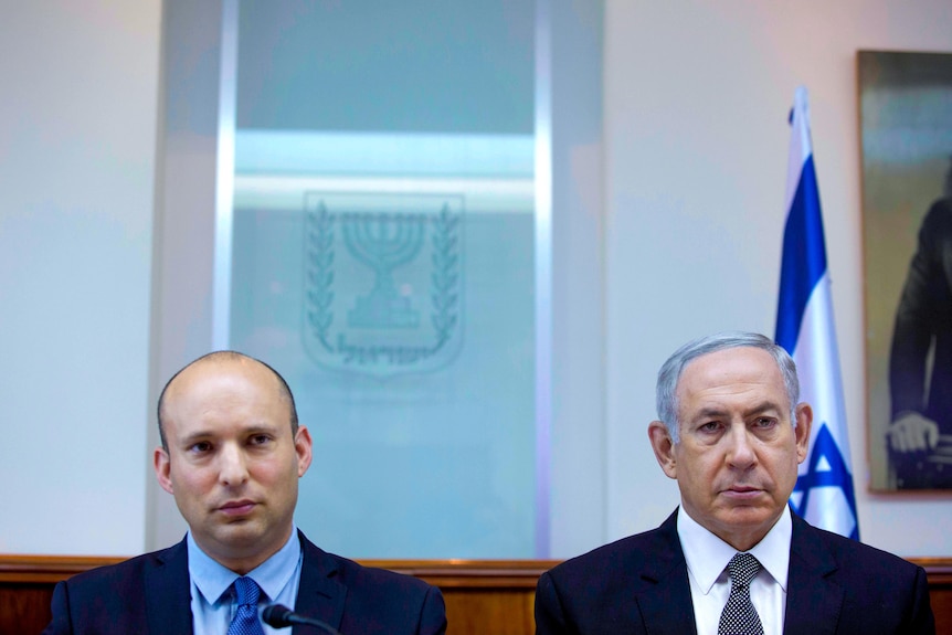 Benjamin Netanyahu and Naftali Bennett sitting stony faced in a room with an Israeli flag behind them