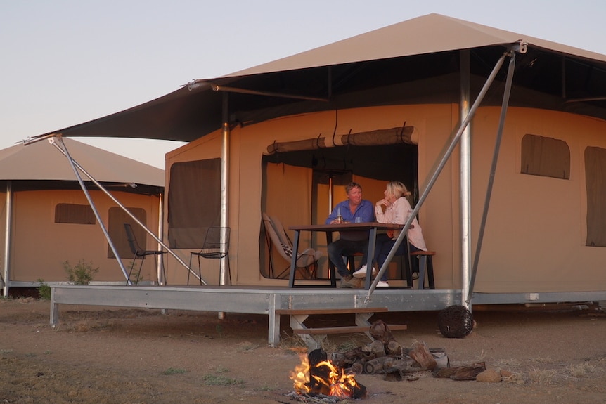 Man and women sit on deck at sunset with beer and wine. A large glamping tent is behind them and small fire in front of them