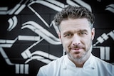 Head shot of MasterChef Jock Zonfrillo, who is standing in front of a large pile of chefs' aprons.