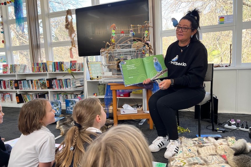A woman with a budgie sitting on her glasses, reading a book to school children sitting on the floor.