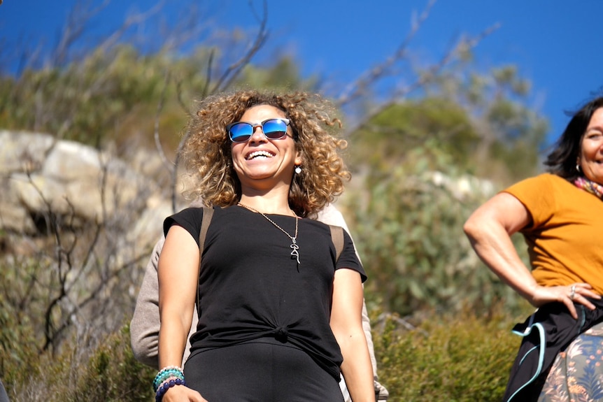 Reem Tawfik wearing blue sunglasses, black shirt, smiling, blue sky and trees behind.