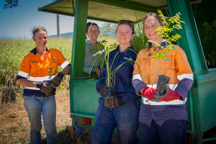 Four women dressed in work clothes stand around a trailer full of sapling trees and plants.