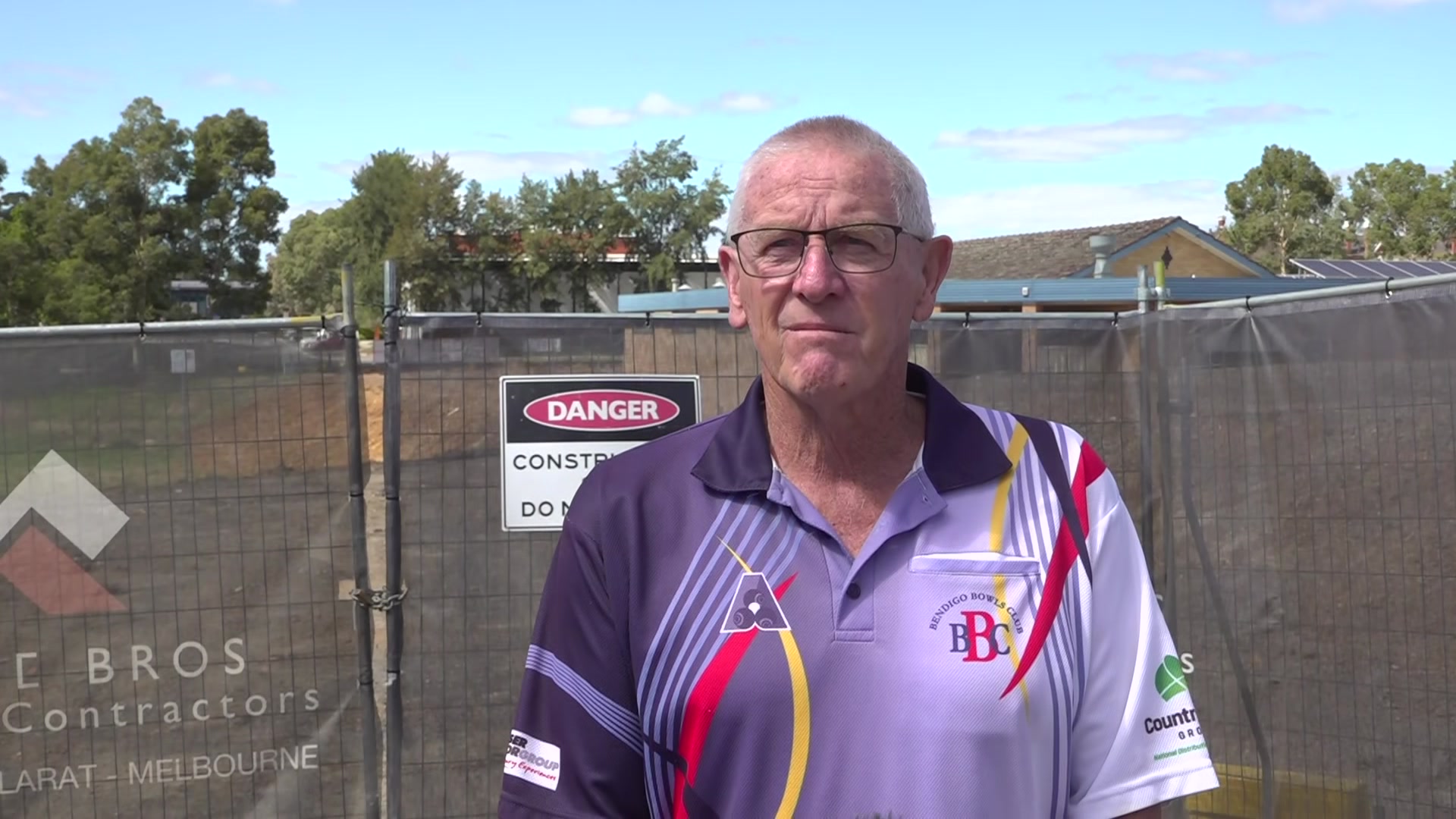 Homem vestindo camisa pólo roxa, branca, vermelha e amarela, óculos, em frente ao portão de um canteiro de obras.