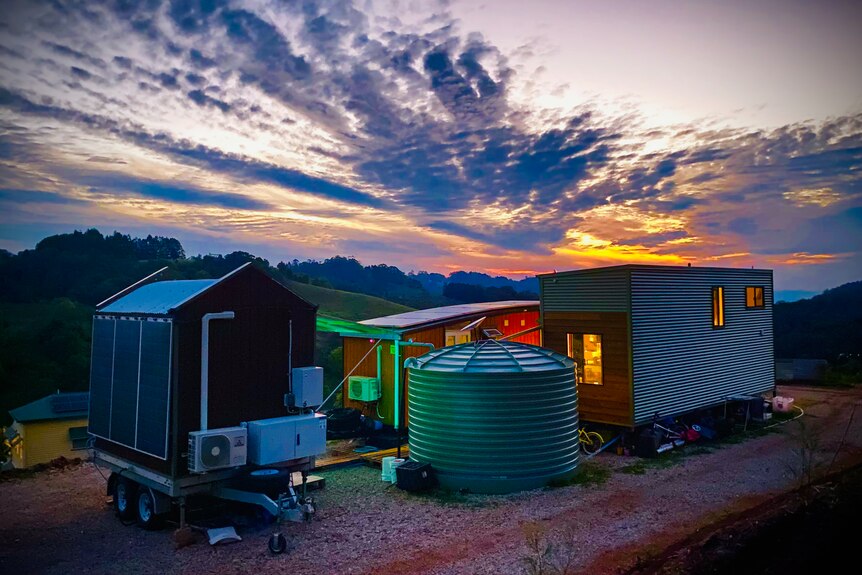 A cluster of three tiny houses stationed around a green water tank, with sunset in the background.