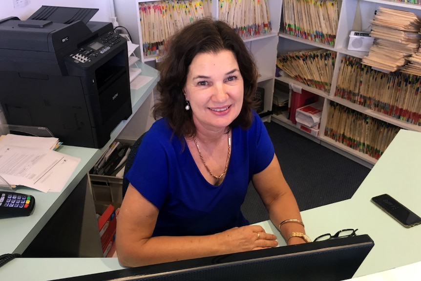 Woman sitting at a desk with a lot of folders in the background. She is smiling.