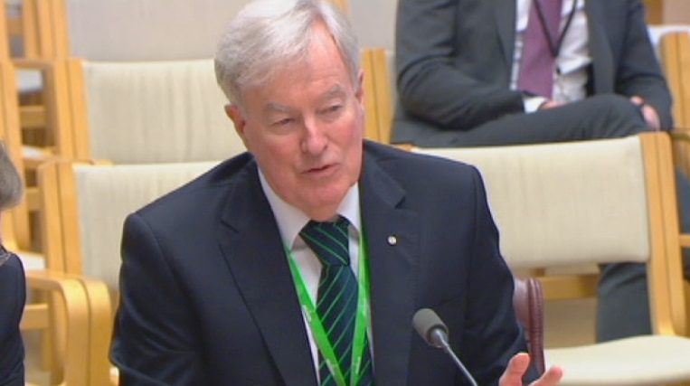 John Lloyd appears before a Senate hearing, answering questions. He's wearing a green lanyard.