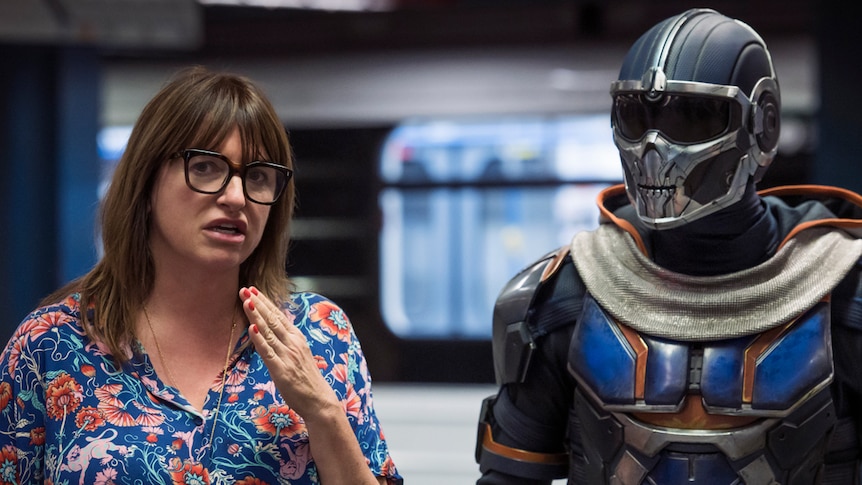 A woman in her early 50s in floral top with glasses directs someone in soldier/robot outfit