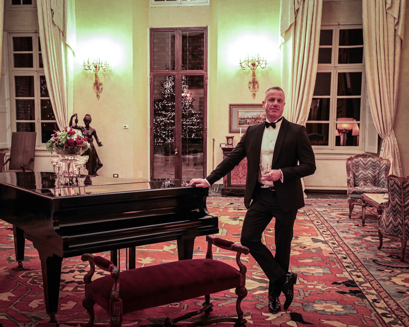 In an opulent room with ornate chairs, grand piano and golden candelabras, smiling man in black suit leans against piano.