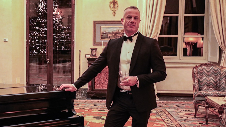 In an opulent room with ornate chairs, grand piano and golden candelabras, smiling man in black suit leans against piano.