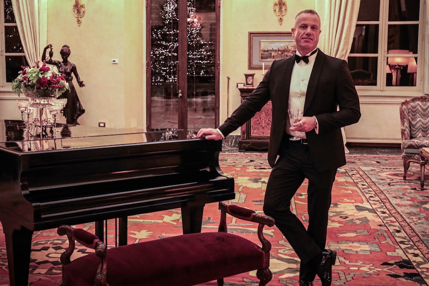 In an opulent room with ornate chairs, grand piano and golden candelabras, smiling man in black suit leans against piano.