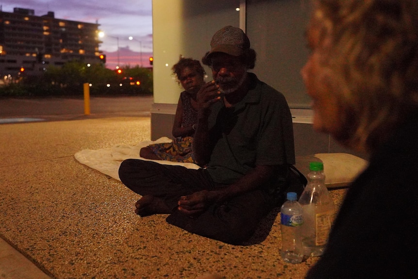 Three Indigenous people sit on the side of a street at dusk