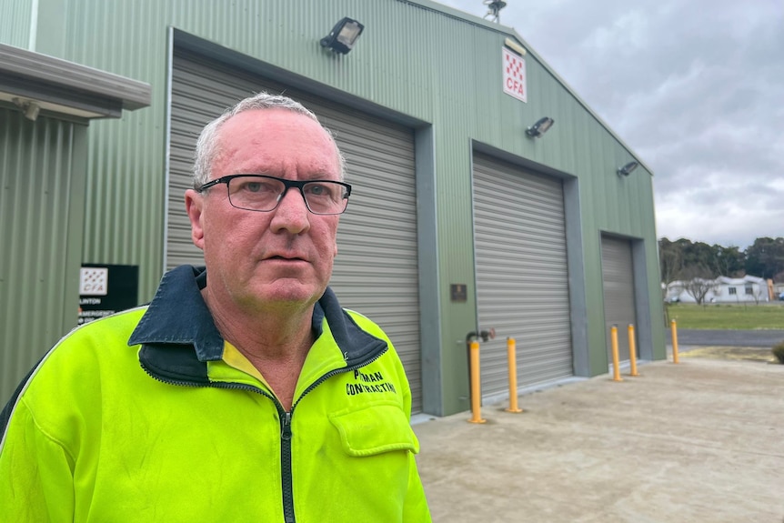 An older man, wears glasses, in a fluoro green jacket looks seriously at the camera with a cfa shed in the background.