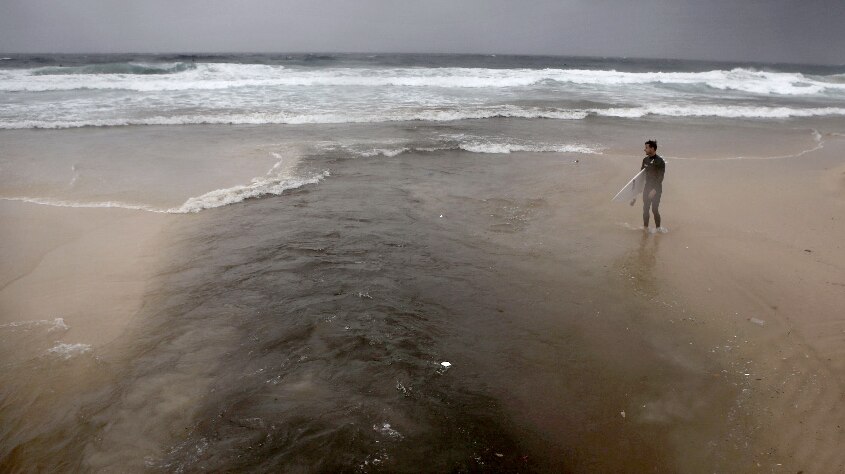 A surfer on the beach during a storm, with dark storm water gushing from drains into the ocean.