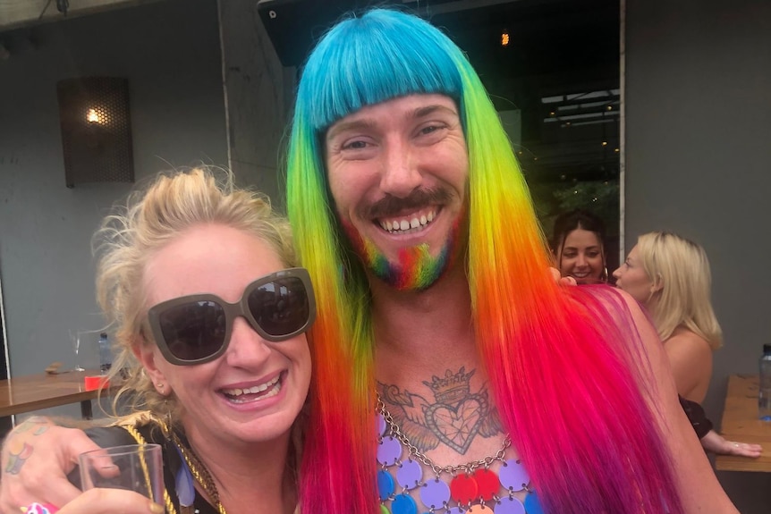 A blonde woman is standing next to a person with a rainbow wig and a rainbow outfit.