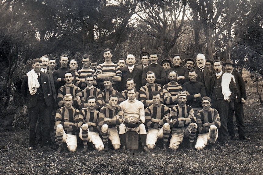 An old black and white photograph of a football team of men wearing woolen jumpers holding a football that reads 1912