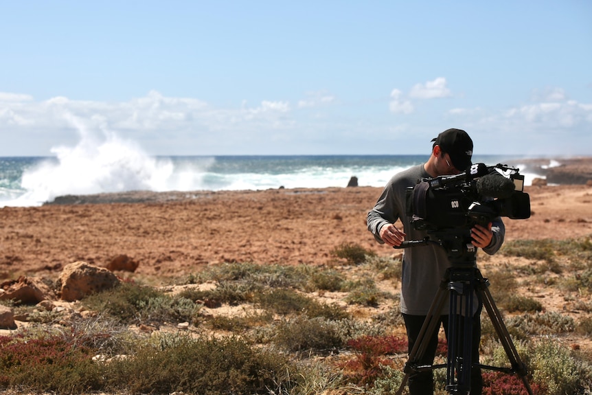 A wave crashed onto rugged cliffs in the background behind an ABC camera operator looking into his viewfinder.