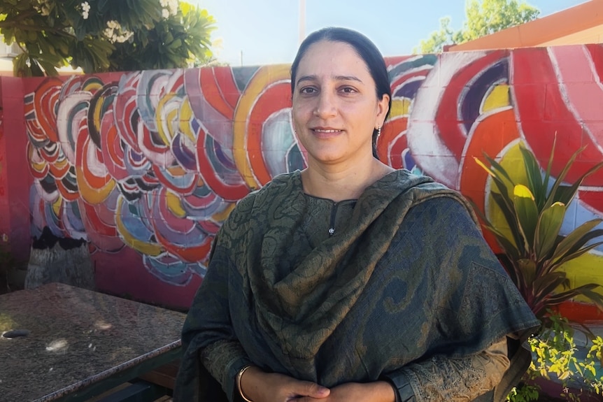 A woman in dark clothing with a colourful wall behind her, wears an Indian-style scarf drapped across both shoulders.